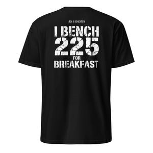 Peut inclure: T-shirt noir avec texte blanc : « I BENCH 225 FOR BREAKFAST ». Le chiffre « 225 » est mis en évidence. Au-dessus du texte, on peut lire « I AM A SPARTAN ». Le texte est en police vieillie.