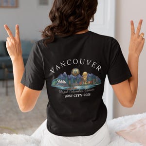 May include: Black t-shirt with a graphic of Vancouver, British Columbia, Canada, featuring mountains, a cityscape, a soccer ball, and a trophy. The text "VANCOUVER" arches across the top, with "HOST CITY 2026" below.