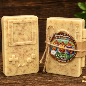 May include: Two bars of handmade soap. One is shaped like a retro handheld game console. The other is a stack of soap bars wrapped with a label that reads "Greylee Farm, Smells Like the 90s, Chicks on the Lawn Goat Milk Soap."