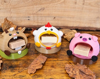 May include: Three animal-shaped soap holders made of plastic. A brown goat, a white chicken, and a pink pig, each with a wide open mouth holding a rectangular bar of soap. The goat sits on a green base, the chicken on a tan base, and the pig on a brown base.
