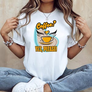 Peut inclure: T-shirt blanc avec un motif sur le thème du café. Le motif présente une tasse de café jaune avec des ailes, un cœur et le texte "Coffee? YES, PLEASE!" en jaune et noir. Le t-shirt est un vêtement décontracté de tous les jours.