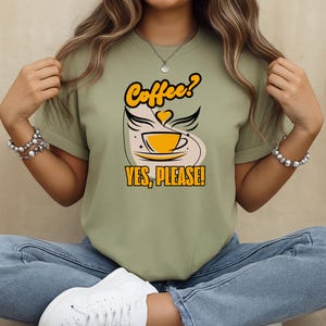 Puede incluir: Camiseta verde claro con un diseño temático de café. El diseño presenta una taza de café amarilla con alas, la palabra "Coffee?" arriba y "YES, PLEASE!" abajo. La camiseta está hecha de un material suave.