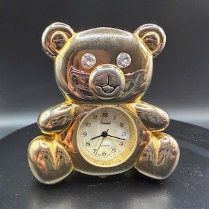 May include: A gold-colored teddy bear-shaped clock with a quartz movement. The clock face is white with black numerals and hands. The bear has crystal accents for eyes and the brand name "Perini" is visible on the clock face.