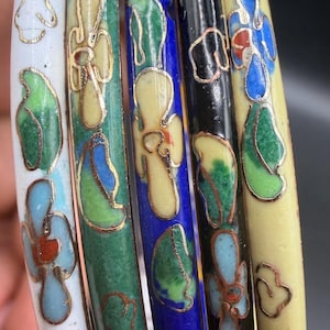 May include: Close-up of several colorful enamel bracelets. The bracelets feature floral designs in white, green, blue, black, and yellow, with gold-colored metal accents. The bracelets are stacked together.