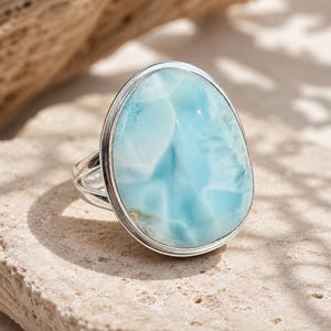 May include: A silver ring featuring a large, oval-shaped, polished Larimar gemstone. The stone displays a marbled pattern of sky blue and white tones. The metal band is smooth and polished, securely holding the stone in a simple bezel setting.