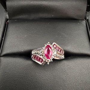 May include: A silver ring with a marquise-cut ruby center stone, surrounded by smaller diamonds and rectangular rubies. The ring is set in a white metal and is displayed in a black jewelry box.