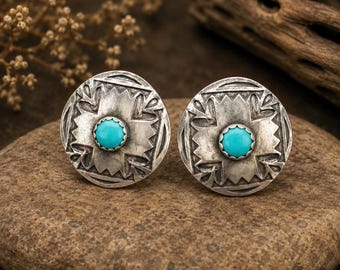 May include: A pair of silver-toned stud earrings with a turquoise center stone. The earrings have a circular design with a cross-like pattern and decorative edges. The earrings are displayed on a stone surface.