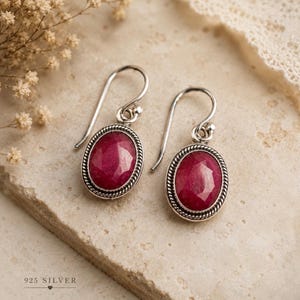 May include: A pair of silver earrings with oval, deep red gemstones. The gemstones are set in a silver frame with a twisted rope design. The earrings have a hook closure. The text "925 SILVER" is at the bottom.
