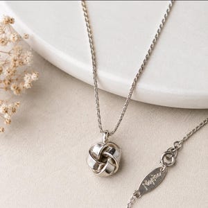 May include: A silver-toned necklace featuring a spherical knot pendant with a polished finish. The chain is a fine, textured link style. A small, oval-shaped metal tag is attached near the clasp, clearly engraved with the brand name Napier.