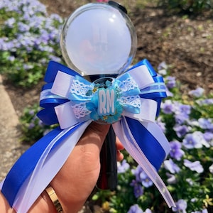 May include: A black and white light stick with a clear orb featuring the BTS logo. The light stick is decorated with blue and white ribbons and a blue flower with the letters "RM".