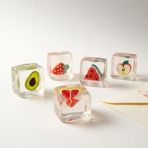 May include: Five clear, cube-shaped resin paperweights, each containing a miniature fruit design. The designs include a green avocado, a red strawberry, a red watermelon slice, a red apple slice, and a red citrus wedge, all suspended in clear material.