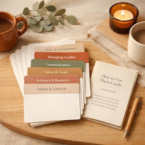 May include: A stack of relationship-themed cards with titles like "Trust & Commitment" and "Family & Lifestyle" sits on a wooden surface. A small booklet titled "How to Use These Cards" and a pen are next to the cards. A lit candle and a mug are also in the image.