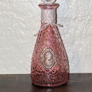 May include: Pink glass perfume bottle with a textured surface and a clear, faceted stopper. The bottle features a cameo design and is wrapped with twine. A decorative item for display.