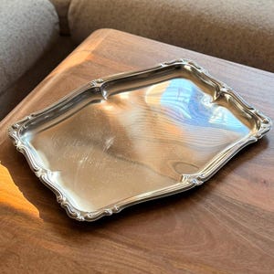Puede incluir: Una bandeja de servicio rectangular de tono plateado con una superficie pulida y reflectante y bordes festoneados decorativos. La bandeja presenta un área central lisa y plana y bordes elevados y ornamentados que se curvan hacia afuera.