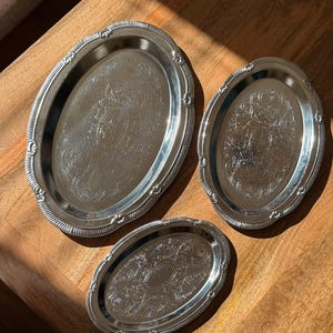 Op de afbeelding: Een set van drie ovale, zilverkleurige metalen dienbladen met ingewikkelde, in reliëf aangebrachte bloemmotieven langs de randen en op de centrale oppervlakken. Elk dienblad heeft een gepolijste, reflecterende afwerking met een geschulpte rand.