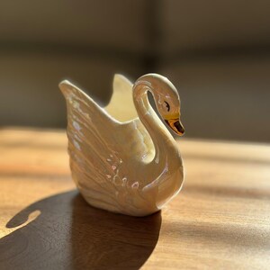 May include: A ceramic swan-shaped planter or decorative vessel featuring an iridescent, pearlescent finish. The swan has a curved neck, detailed moulded wing feathers, and a gold-painted beak. The interior is hollow and finished in white.