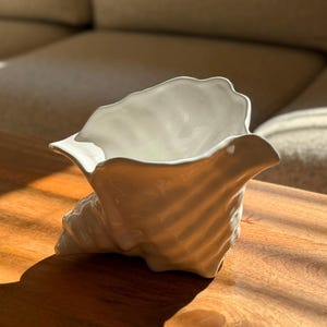 Op de afbeelding: Een witte decoratieve schaal van keramiek in de vorm van een schelp. Het object heeft een glanzende, gladde afwerking met organische, golvende ribbels en een brede opening, die het licht reflecteert op de gebogen, sculpturale vorm.