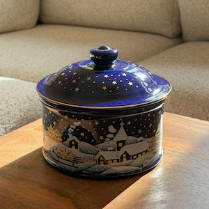 Op de afbeelding: Een ronde, donkerblauwe keramische pot met deksel, voorzien van een goudkleurige winterdorp-scène met besneeuwde huizen, bomen en een sterrenhemel. De rand van het deksel en de basis zijn afgewerkt met een dunne gouden lijn.