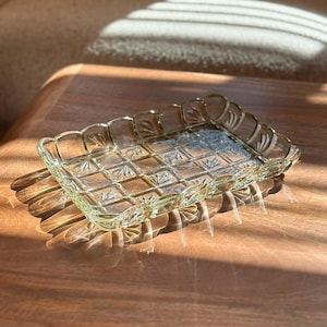 Könnte beinhalten: Ein rechteckiges Tablett aus klarem Glas mit einem Gittermuster auf der Unterseite und einem gewellten Rand mit geprägten blattartigen Motiven. Das Glas hat einen dezenten hellgrünen Schimmer und eine reflektierende Oberfläche.