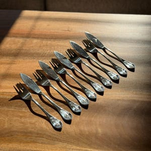 May include: A set of eight small silver-toned metal utensils, consisting of four forks and four knives arranged in an alternating diagonal row. Each piece features a slender handle with a decorative embossed pattern at the base and a polished finish.