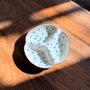Op de afbeelding: Een ronde, gebroken witte keramische schaal met drie aparte vakken. Het binnenoppervlak is versierd met een herhalend patroon van kleine blauwe bloemblaadjes en piepkleine oranje stippen die over de gehele bodem en wanden zijn verspreid.