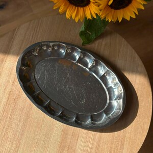 May include: An oval-shaped silver-colored metal tray featuring a scalloped edge design and a smooth, reflective center surface with visible light surface scratches. The tray has a metallic finish and a raised, fluted rim around the perimeter.
