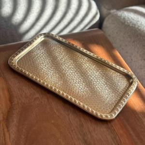 Pode incluir: Uma bandeja retangular de metal dourado com uma superfície texturizada e martelada no centro e uma borda elevada decorada com um padrão repetitivo de pequenas contas circulares espaçadas uniformemente ao longo de toda a borda externa.