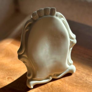 Può includere: Una cornice portafoto in ceramica color bianco sporco lucido con un centro ovale e un bordo decorativo smerlato. La cornice presenta curve fluide in rilievo lungo i bordi e una superficie liscia e riflettente che cattura la luce.