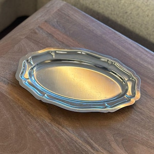 Puede incluir: Una bandeja de servicio ovalada de metal plateado con una superficie pulida y reflectante y un borde decorativo festoneado. La bandeja presenta una base central lisa y plana rodeada por un borde elevado y contorneado con muescas.