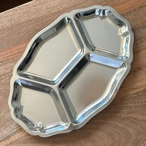 Op de afbeelding: Een gepolijst zilverkleurig metalen serveerblad met een geschulpte rand en een verdeelde binnenkant met drie aparte compartimenten. Het blad heeft een reflecterend, glad oppervlak en een langwerpige, afgeronde rechthoekige vorm.