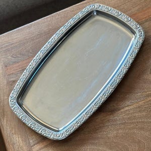 Può includere: Un vassoio rettangolare in metallo color argento con una superficie centrale liscia e piatta e un bordo rialzato decorato con un intricato motivo a volute in rilievo che si ripete lungo tutto il bordo esterno della cornice.