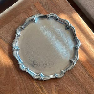 Peut inclure: Un plateau rond en métal argenté doté d'un bord festonné et d'un motif gravé décoratif au centre. Le plateau présente une finition polie avec une surface réfléchissante et un rebord surélevé qui suit le design incurvé et orné de la bordure.