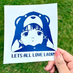 May include: A blue and white sticker featuring an anime-style character wearing a bear hat. The sticker has the text "LETS ALL LOVE LAIN!" in a digital font. The sticker is held against a green grassy background.