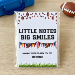 May include: A rectangular card in a clear plastic sleeve featuring the text "LITTLE NOTES BIG SMILES" and "LUNCHBOX NOTES FOR GAME DAY AND JUST BECAUSE". The design includes brown football illustrations, a colorful pennant banner, and green grass.