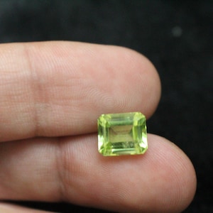 May include: A square-cut, light green gemstone with a faceted surface. The gem is held between fingers against a dark background, showcasing its clarity and colour. The gemstone's edges are sharp and well-defined.
