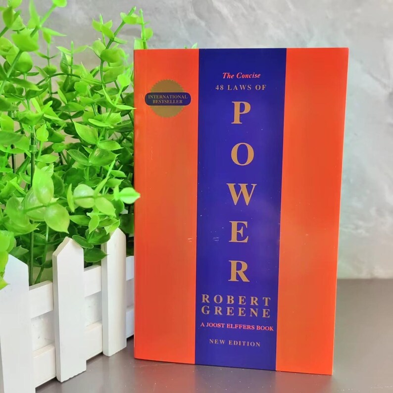 Op de afbeelding: Een hardcover boek getiteld "The Concise 48 Laws of Power" van Robert Greene. Het boek heeft een oranje omslag met een verticale blauwe streep in het midden. De titel staat in grote witte letters. Nieuwe editie, internationale bestseller.