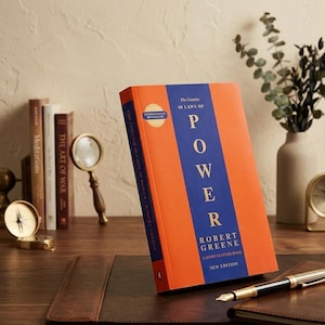 Op de afbeelding: Een oranje en blauw boek getiteld "The Concise 48 Laws of Power" van Robert Greene. Het boek staat op een bureau met een pen, een vergrootglas en andere boeken op de achtergrond. Een kompas en een klok zijn ook zichtbaar.