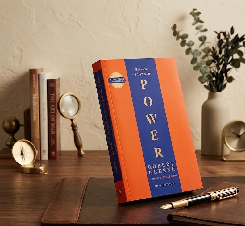 Op de afbeelding: Een oranje en blauw boek getiteld "The Concise 48 Laws of Power" van Robert Greene. Het boek staat op een bureau met een pen, een vergrootglas en andere boeken op de achtergrond. Een kompas en een klok zijn ook zichtbaar.