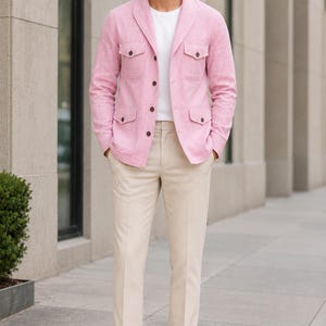 May include: A light pink textured jacket with a shawl collar, four front pockets with dark buttons, and a central button closure. The jacket is worn over a plain white t-shirt, paired with beige trousers and brown leather loafers.