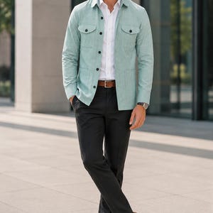 Pode incluir: Uma sobrecamisa de manga comprida em verde menta claro com dois bolsos no peito, usada sobre uma camisa branca de colarinho. O conjunto inclui calças pretas, um cinto de couro castanho com fivela prateada e sapatos de couro castanhos.
