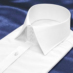 Puede incluir: Una camisa de vestir blanca y nítida con un cuello alto y rígido que presenta cuatro botones verticales en la banda del cuello. La camisa tiene un diseño de cuello puntiagudo estructurado y un solo botón visible en la tapeta frontal.