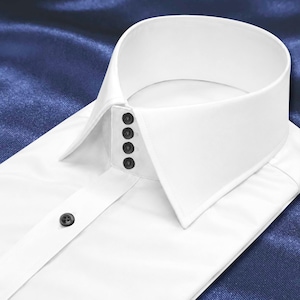 Puede incluir: Una camisa de vestir blanca y nítida con un cuello alto y estructurado que presenta cuatro botones negros dispuestos verticalmente a lo largo de la banda del cuello. Un botón negro individual es visible en la tapeta frontal de la camisa.