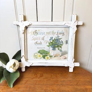 May include: A rectangular wall sign in a distressed white wooden frame with butterfly corner accents. The center features a floral illustration with blue and yellow flowers and the text Grieve not the Holy Spirit of God. Ephesians 4:30.