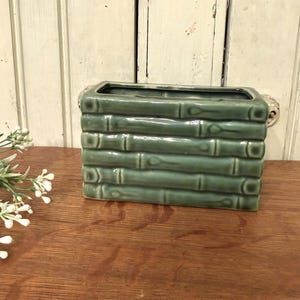 May include: A rectangular, green ceramic vase with a bamboo design. The vase is a muted green color and has a glossy finish. The bamboo design is detailed and adds texture to the vase. The vase is sitting on a wooden surface.