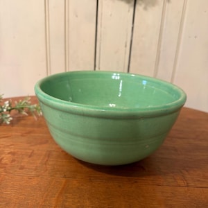 May include: A vintage, light green ceramic mixing bowl. The bowl has a rounded shape with a slightly flared rim and horizontal ridges around the exterior. The bowl is sitting on a wooden surface.