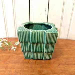 May include: A square, green ceramic planter with a textured, stacked design. The planter has a wide opening and is set on a wooden surface. The color is a vibrant teal, and the design is reminiscent of a woven basket.