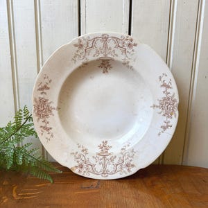 May include: A white antique plate with a brown floral design around the rim and center. The plate has a scalloped edge and is set on a wooden surface. The plate is a vintage piece.