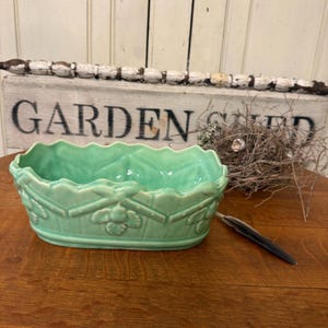 May include: A rectangular, light green ceramic planter with a scalloped rim and floral relief design. The planter sits on a wooden surface, with a weathered sign that reads "GARDEN SHED" in the background.