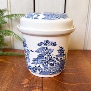 May include: A white ceramic butter dish with a blue Willow pattern design. The dish has a lid and features a detailed landscape scene with trees, buildings, and a bridge. The design is repeated on the lid.