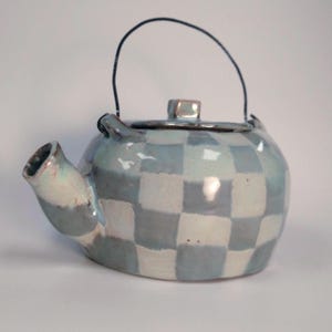 May include: A ceramic teapot featuring a light blue and white checkered pattern across its rounded body. The teapot includes a matching lid with a small knob, a short spout, and a thin, dark metal wire handle attached to two small loops.